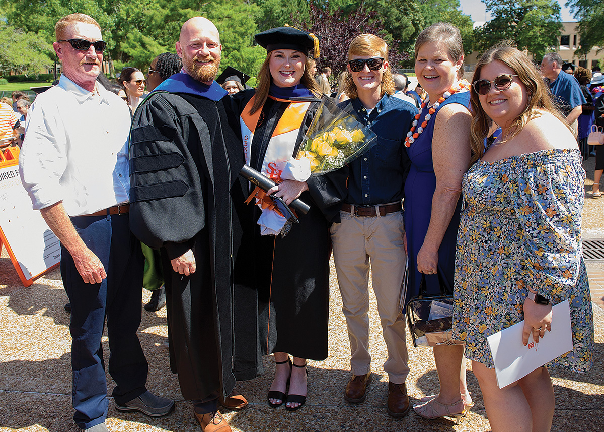 A graduate, their professor, and the graduate's family all pose outsdie of the Johnson Coliseum on graduation day. The graduate has a First Generation sash and is holding a bouquet of flowers.