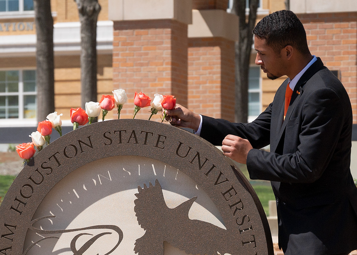 A college student in a suit gently places a rose in the Raven's Call structure at the foot of the clocktower in memory of an alumni who has passed.